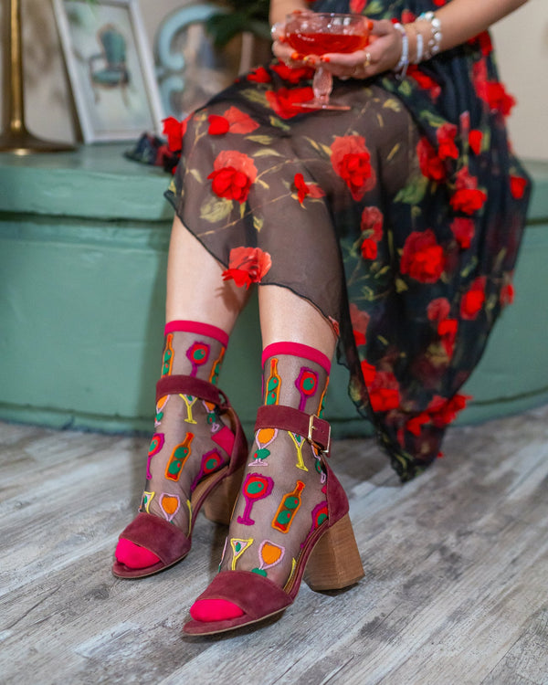 Person wearing a floral dress and colorful shoes and socks with cocktails on it with a glass of red drink in hand.