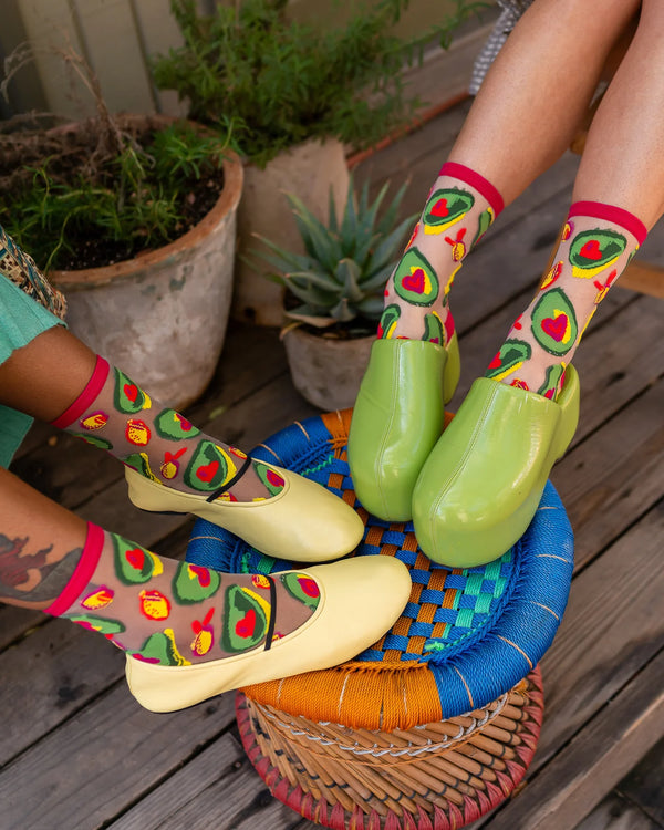 Person wearing colorful socks with avocado and heart patterns and yellow shoes on a wooden deck.
