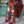 Person wearing a floral dress and colorful shoes and socks with cocktails on it with a glass of red drink in hand.