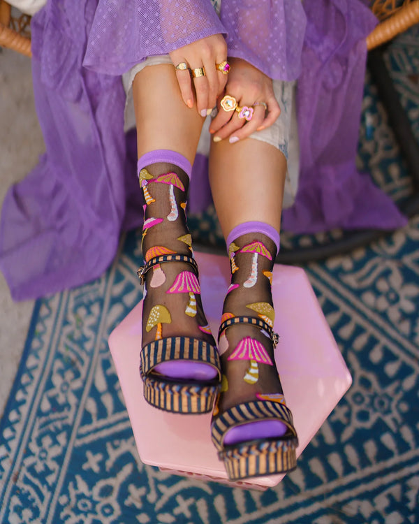Person wearing colorful platform sandals with a colorful mushroom patterned sock and purple shoe, sitting on a pink stool.
