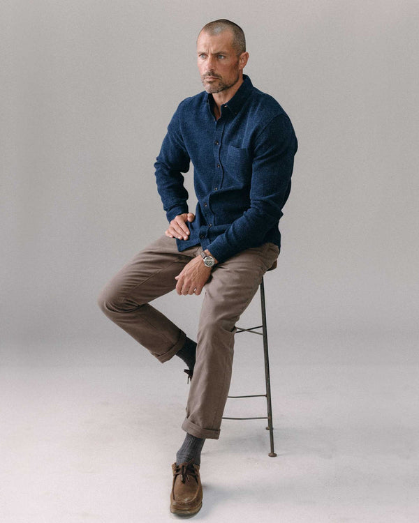 Man sitting on a stool wearing a blue sweater and brown pants against a plain background