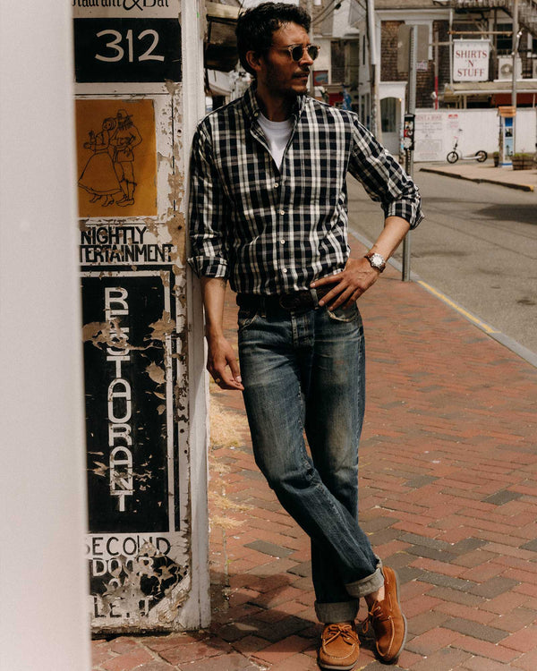 Man in plaid shirt and jeans leaning against a wall on a street.