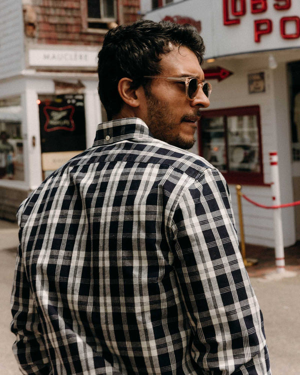 Man wearing a dark navy and white plaid shirt with sunglasses, standing in front of a vintage-style building.