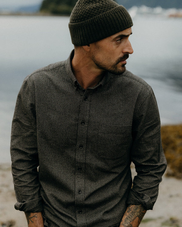 Man wearing a dark knit beanie and button-up shirt standing by a body of water.